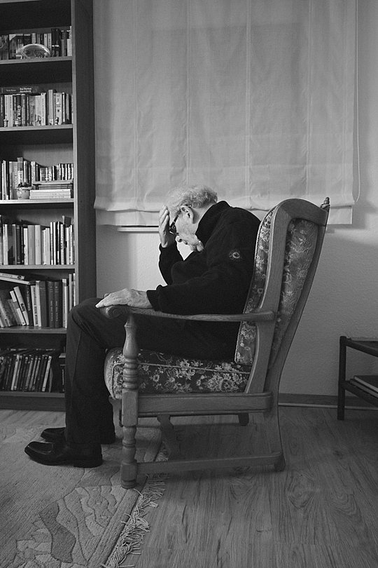 Älterer Mann sitzt nachdenklich in einem Sessel vor einem Bücherregal.