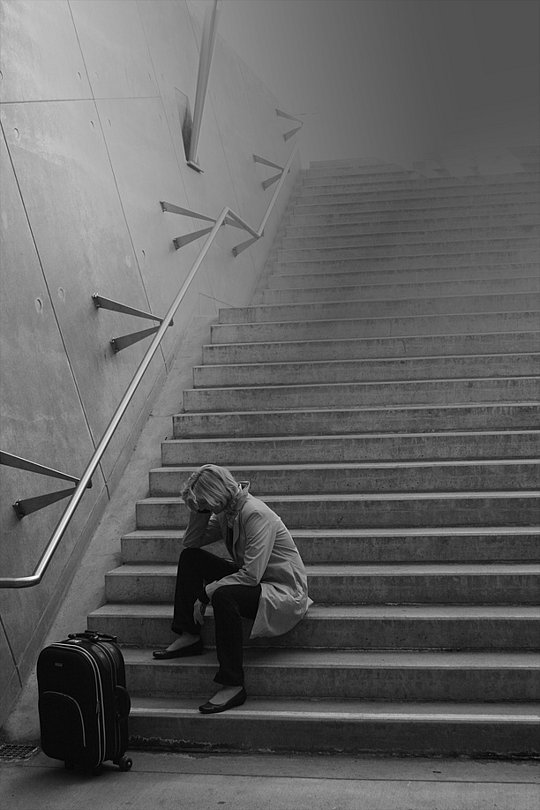 Frau sitzt erschöpft auf einer Treppe neben einem Koffer.