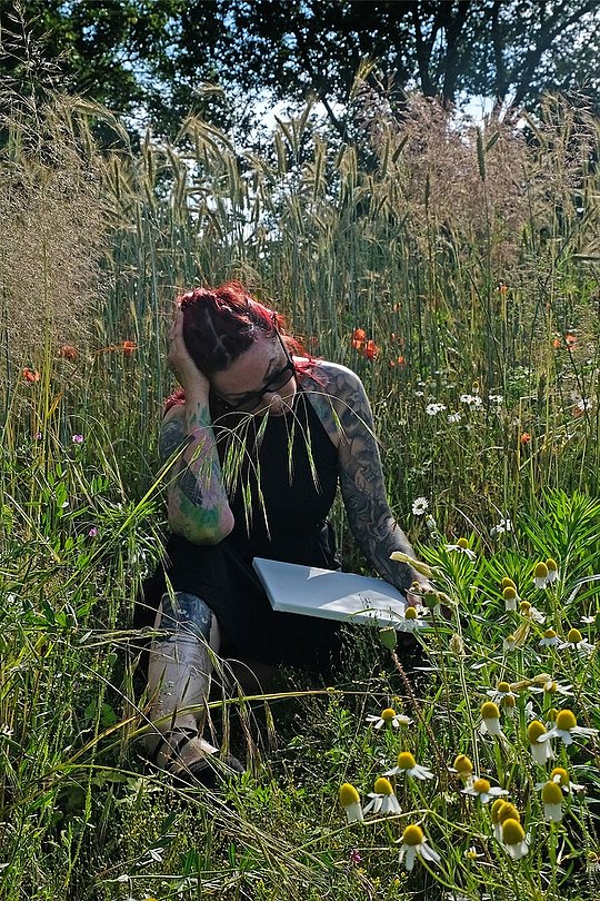 Person sitzt nachdenklich in einer Wiese mit Blumen und Notizbuch.