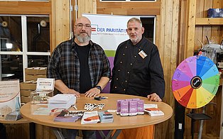 Zwei Männer an einem Infostand mit Infomaterial, Spendenbox und einem bunten Glücksrad.