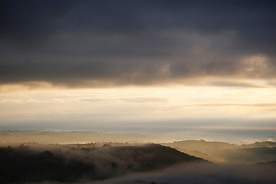 Nebel liegt über hügeliger Landschaft unter dunklen Wolken.